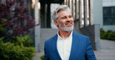 Gülümseyen ve başarılı bir iş adamı portresi, modern ofis binasının önünde sokakta yürürken, özgüvenle şirket yöneticisinin kamera vesikalığına bakıyor.