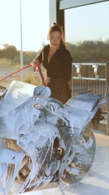 25 yaşında, koyu renk saçlı, şık giyimli, yüksek atkuyruklu, lüks demir atı yıkayan, yüksek basınçlı jet akımında köpüklü hortumla araba yıkama hizmeti veren, şık bir kız-motosikletçi.