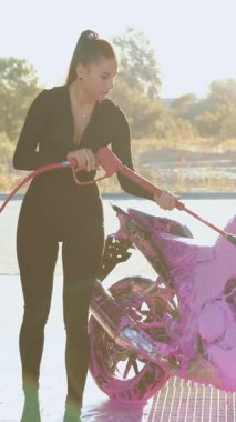 Sevimli, çekici, 25 yaşında, at kuyruklu, siyah saçlı esmer, açık hava oto yıkama istasyonunda pembe köpüklü spor bir motosiklet giyiyor. Kapatın.