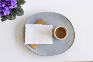 Bahar kırtasiyesi sahnesi. Boş tebrik kartı, toprak kaplama tabağı, bir fincan kahve, Afrika menekşe çiçeği ya da menekşe azizi çiçekleri leylağı. Beyaz masa arkaplanı, düz yatak, üst manzara. Yiyecek konsepti.