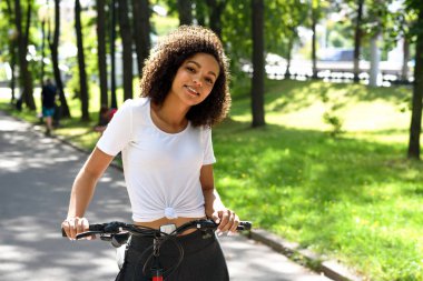  Parkta bisiklete binen neşeli Afro-Amerikan kadın. Çekici, zayıf bir kız, sağlıklı bir hayat sürüyor..