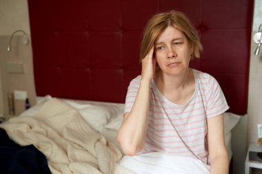 Orta yaşlı bir kadın yatakta oturuyor, başını tutuyor. Sağlık sorunları, baş ağrısı, menopoz, uykusuzluk.