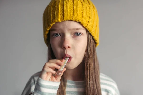 Retrato de uma menina triste infeliz em um chapéu, nariz escorrendo e dor de cabeça, mede a temperatura com um termômetro. — Fotografia de Stock