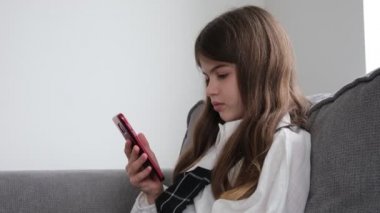Okul üniformalı küçük, genç, güzel bir kız elinde telefonla kanepede oturuyor.