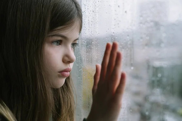 Pequena triste, menina olha pela janela, fora da janela está chovendo — Fotografia de Stock