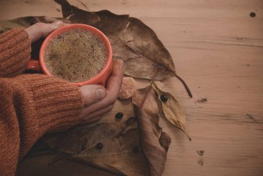 Erkek elleri sıcak kahve kapuçinosu, turuncu süveter ve eski ahşap arka planda sonbahar yaprakları tutuyor. Antika tonlar. Sonbahar rahatlama konsepti. Boşluğu kopyala