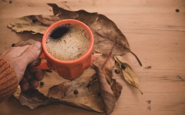 Erkek eli sıcak kahve kapuçinosu, turuncu kazak ve eski ahşap arka planda sonbahar yaprakları tutuyor. Antika tonlar. Sonbahar rahatlama konsepti. Boşluğu kopyala