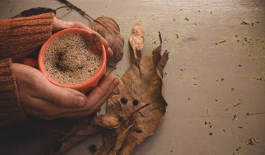 Erkek elleri sıcak kahve kapuçinosu, turuncu süveter ve eski gri ahşap arka planda sonbahar yaprakları tutuyor. Antika tonlar. Sonbahar rahatlama konsepti. Boşluğu kopyala