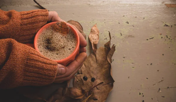 Erkek elleri sıcak kahve kapuçinosu, turuncu süveter ve eski gri ahşap arka planda sonbahar yaprakları tutuyor. Antika tonlar. Sonbahar rahatlama konsepti. Boşluğu kopyala