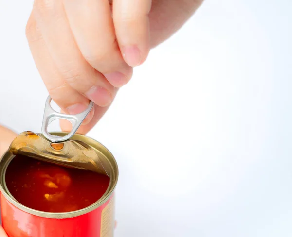 Woman's hand opens lid of canned fish on white background. Sardine in tomato sauce. Processed food. Canned fish in tin can. Seafood in can container. Preserve food. Tin can for storage preserve food.