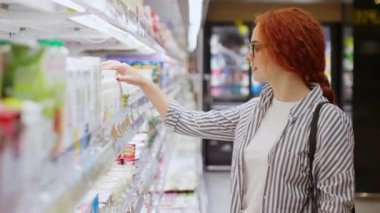 Sıradan giyinmiş, gözlüklü çekici kızıl saçlı kız modern markette özenle yoğurt seçiyor.