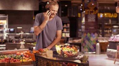 Arabası ürünlerle dolu yakışıklı sarışın sakallı adam modern markette karısıyla telefonda konuşuyor.