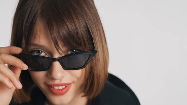 Şık mavi gözlü, kısa saçlı, kırmızı dudaklı güneş gözlüklü, beyaz arka planda izole edilmiş kameraya güvenle bakan bir kız.