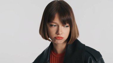 Sıkılmış, esmer, kısa saçlı, sıradan giyinmiş, beyaz arka planda ilgisiz poz veren bir kız. Üzücü ifade