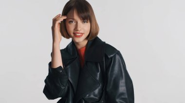 Kırmızı dudaklı, deri ceketli, güzel, sırıtan esmer kız beyaz arka planda izole edilmiş kameraya bakıyor. Moda kızı.