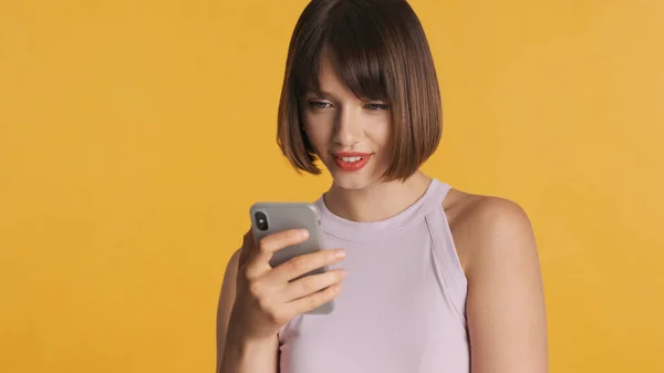 Pretty brunette girl with bob hair chatting with friends online on smartphone over yellow background. Always in touch