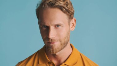 Yakışıklı, sarışın, sakallı adam mavi arka planda kameraya bakıyor.