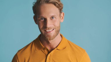 Çekici sarışın sakallı adam kameraya bakıyor ve mavi arka planda izole bir şekilde gülümsüyor. Yüz ifadesi