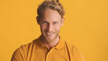 Sarışın, yakışıklı, sakallı bir adam. Kamerada neşeli poz veriyor. Sarı arka planda.