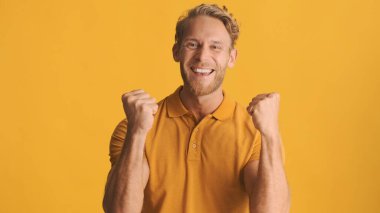 Genç, çekici, sarışın, sakallı adam heyecanlı görünüyor. Renkli arka planda kameraya evet işareti yapıyor. Neşeli ifade
