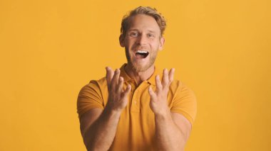 Heyecanlı sarışın sakallı genç adam, sarı arka planda izole edilmiş kamerada duygusal olarak seviniyor. Aşırı neşeli ifade