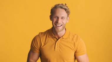 Sarışın, çekici ve sakallı bir adam sarı arka planda kameraya seviniyor. Aşırı neşeli ifade