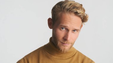 Sıradan giyinmiş yakışıklı sarışın adam beyaz arka planda kameraya dikkatlice bakıyor.