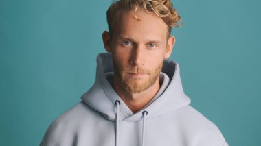 Kapüşonlu sarışın yakışıklı adam mavi arka planda kendine güveniyor. Stüdyoda poz veren erkek model.