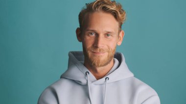 Çekici, şık, sarışın, kapüşonlu sakallı adam kameraya bakıyor ve mavi arka planda tek başına gülümsüyor.