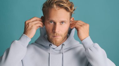 Çekici sarışın sakallı adam sıradan bir kapüşonlu giyip renkli arka planda kablosuz kulaklık takıyor.