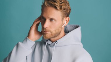Kapüşonlu sakallı yakışıklı adam ciddi görünüyor mavi arka planda izole edilmiş kablosuz kulaklıkla konuşuyor. Modern teknoloji