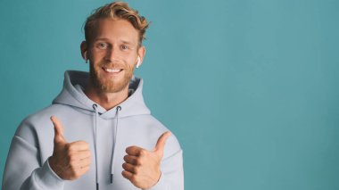 Attractive smiling blond bearded man in hoodie wear wireless earphones keep thumbs up near place for advertisement or promotional text isolated on blue background. Like expression clipart