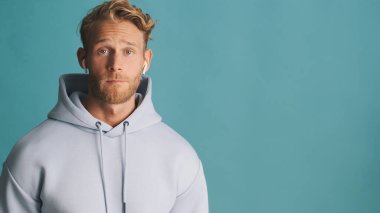 Çekici, şık, sarışın, kapüşonlu sakallı adam reklam için yanında duran kablosuz kulaklıklar takıyor ya da mavi arka planda izole edilmiş promosyon metinleri.