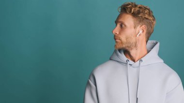 Kapüşonlu sakallı şık sarışın adamın yan görünüşü reklam için yanında duran kablosuz kulaklık takıyor ya da mavi arkaplan üzerinde tanıtım metni takıyor.