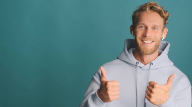 Kapüşonlu sakallı, havalı sarışın adam kablosuz kulaklık takıyor. Reklam ya da renkli arka planda tanıtım metinleri için başparmaklarını havada tutuyor. Aferin ifade.