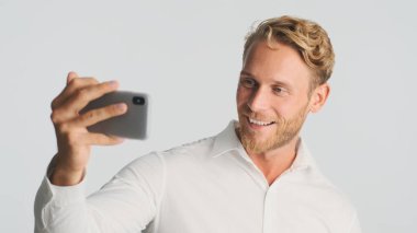 Yakışıklı, sarışın, sakallı iş adamı. Beyaz arka planda akıllı telefondan video görüşmesi yapıyor. Modern teknoloji kavramı