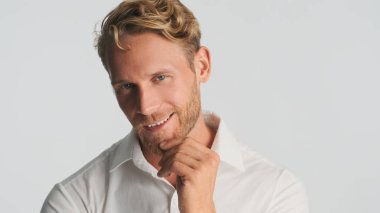 Başarılı sarışın sakallı adam kameraya sinsice bakıyor ve beyaz arka planda izole bir şekilde gülümsüyor.