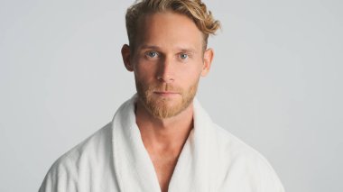 Bornozlu yakışıklı sarışın adam beyaz arka planda izole edilmiş kameraya dikkatlice bakıyor.