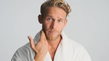 Bornozlu çekici sarışın sakallı adam beyaz arka planda kameraya poz vermeyi düşünüyor.