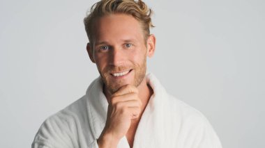 Yakışıklı, sarışın, sakallı, bornozlu adam beyaz arka planda izole edilmiş kamerada mutlu görünüyor.