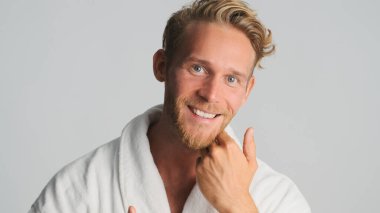 Bornozlu çekici sarışın sakallı adam beyaz arka planda kameraya sevinçle poz veriyor.