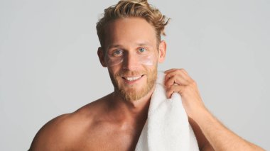 Çekici, sarışın, göz bantlı, omuzlarında havluyla beyaz arka planda mutlu görünen sakallı adam.