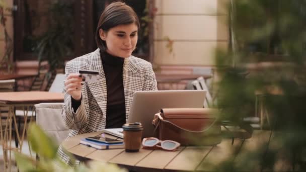 漂亮的微笑女士用信用卡支付网上购物费用 年轻的女商人在户外咖啡馆喝咖啡时在笔记本电脑上付了钱 — 图库视频影像