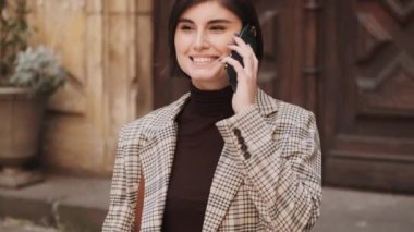 Güzel, şık bir iş kadını, ekoseli siyah giyinmiş, sokakta yürürken telefonla konuşuyor.