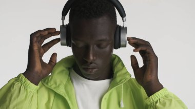 Parlak ceketli yakışıklı Afro-Amerikalı adam beyaz arka planda kulaklıkla müzik dinliyor.