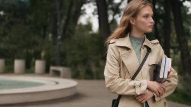 Trençkot giymiş, elinde kitaplarla caddede kamerayla gülümseyen güzel sarışın kız. Üniversiteye giden kız öğrenci.