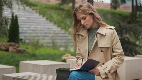 在城市公园的长椅上 穿着风衣的漂亮金发女学生在笔记本上做笔记 — 图库视频影像
