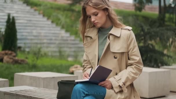 在公园的长椅上 穿着风衣写笔记本的迷人女生 — 图库视频影像