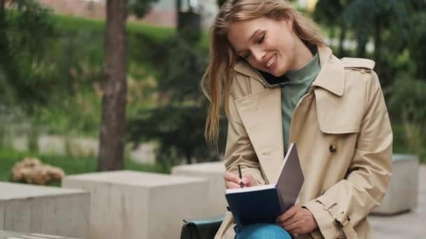 漂亮的女学生看上去很高兴与朋友通电话 并在公园的长椅上的笔记本上记笔记 学习概念 — 图库视频影像