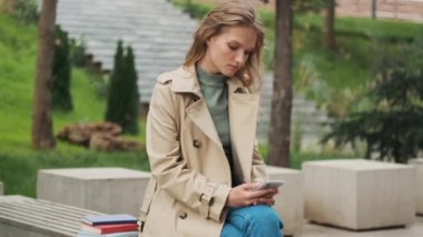 Trençkot giymiş çekici bir öğrenci. Parktaki bankta ders arası sırasında akıllı telefondan arkadaşıyla sohbet ediyor.